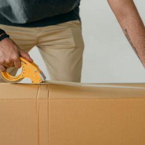 Crop anonymous young male packer in casual clothes using tape gun dispenser for sealing cardboard boxes