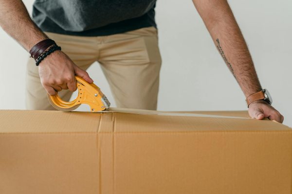 Crop anonymous young male packer in casual clothes using tape gun dispenser for sealing cardboard boxes